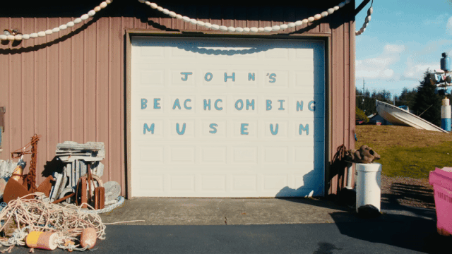 a gif from a short documentary showing a man wearing a black hat and jeans as he walks to the front of a garage door painted with the words "John's Beachcombing Museum"