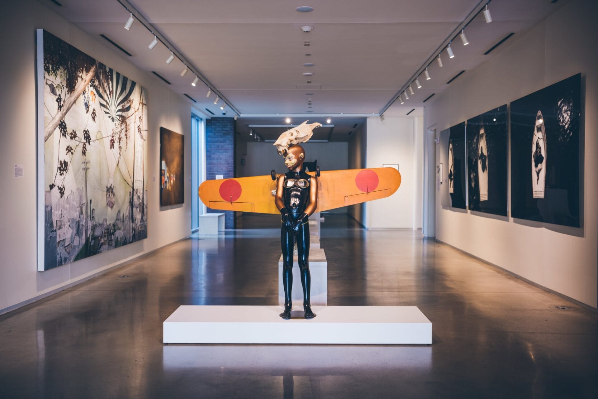 a figurative sculpture with a skull on its head and a yellow pair of wings attached to its back stands in a large illuminated gallery