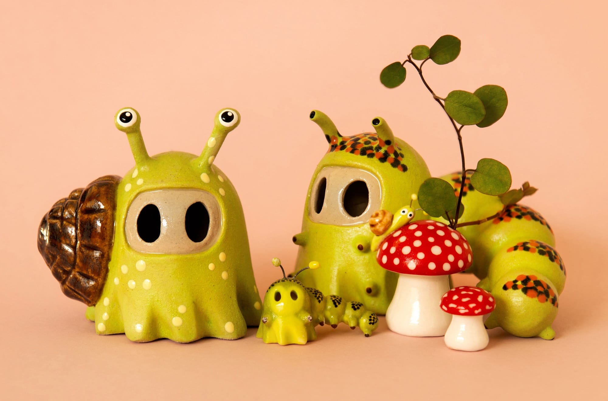 2 small ceramic ghosts posing a green snails and slug. one snail is significantly smaller and rests inbetween the two larger, implying it is a baby. Thet are surrounded by tiny red spotted mushrooms and a sprig with leaves