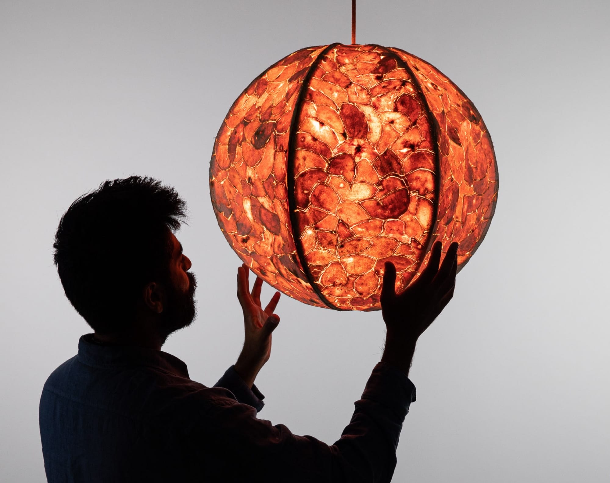 a male figure, silhouetted, reaches out to hold a glowing pendant lamp with a shade made out of sewn-together orange peels