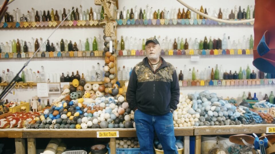 a bearded man with a black sweatshirt, black hat, and jeans, stands in front of a display of hundreds of glass bottles, buoys, and labels in his self-made beachcombing museum