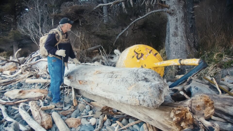 a still from a short documentary showing a man in a black hat and jeans, wearing a backpack and holding a walking stick, as he peers over a giant driftwood log to look at a large yellow buoy washed up onshore