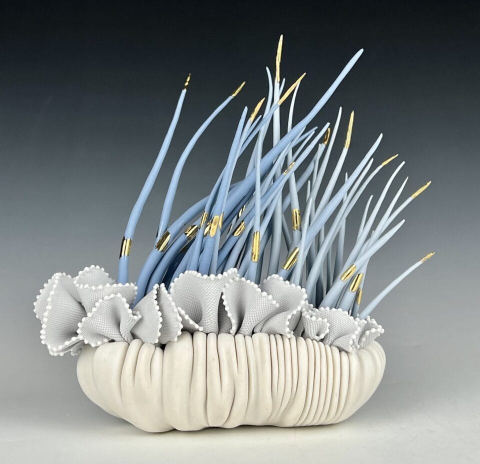 a porcelain sculpture that resembles a bundle of textile with frills, with blue grass-like spikes or fronds emerging from it