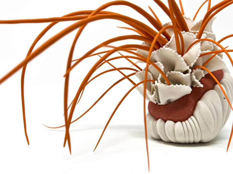 a detail of a porcelain sculpture that resembles a bundle of white-and-red textile with frills, with orange plant-like textured spikes or fronds emerging from it