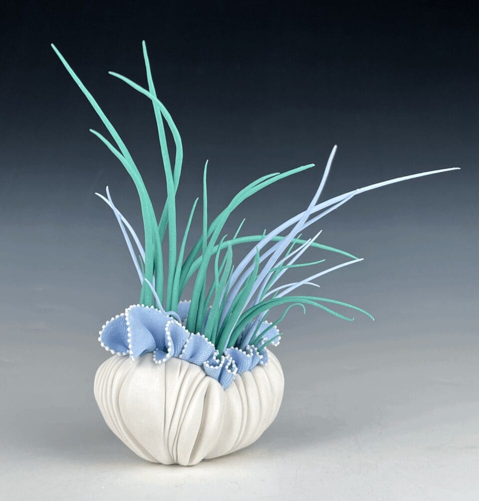 a porcelain sculpture that resembles a bundle of textile with frills, with blue grass-like spikes or fronds emerging from it