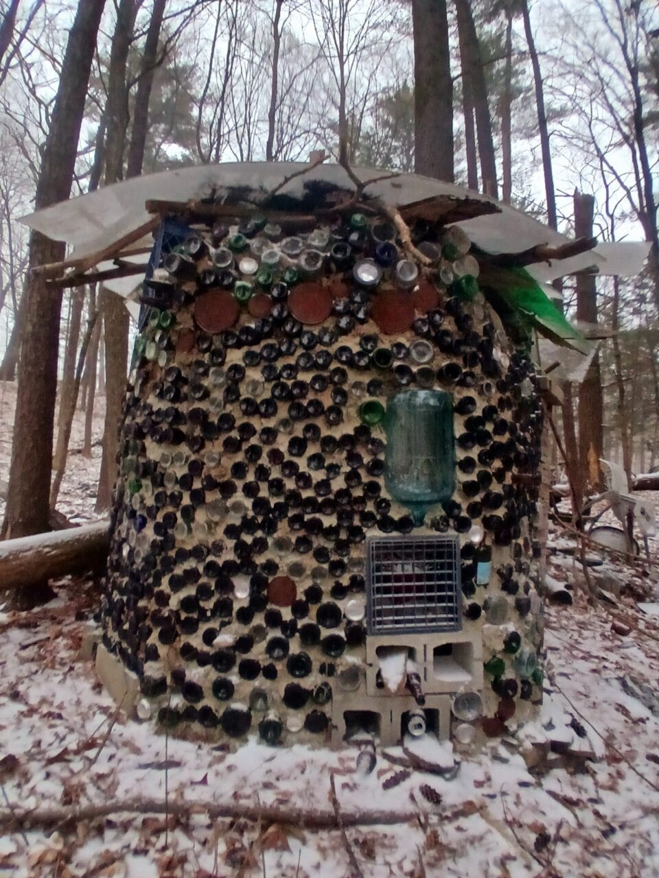 a hand-built cabin or shed-like structure coated in bottles in a snowy wooded landscape, part of "B-Home," a collection of intuitively built structures from salvaged materials