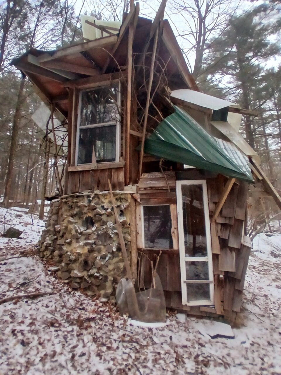 a hand-built cabin or shed-like structure with numerous wonky windows in a snowy wooded landscape, part of "B-Home," a collection of intuitively built structures from salvaged materials