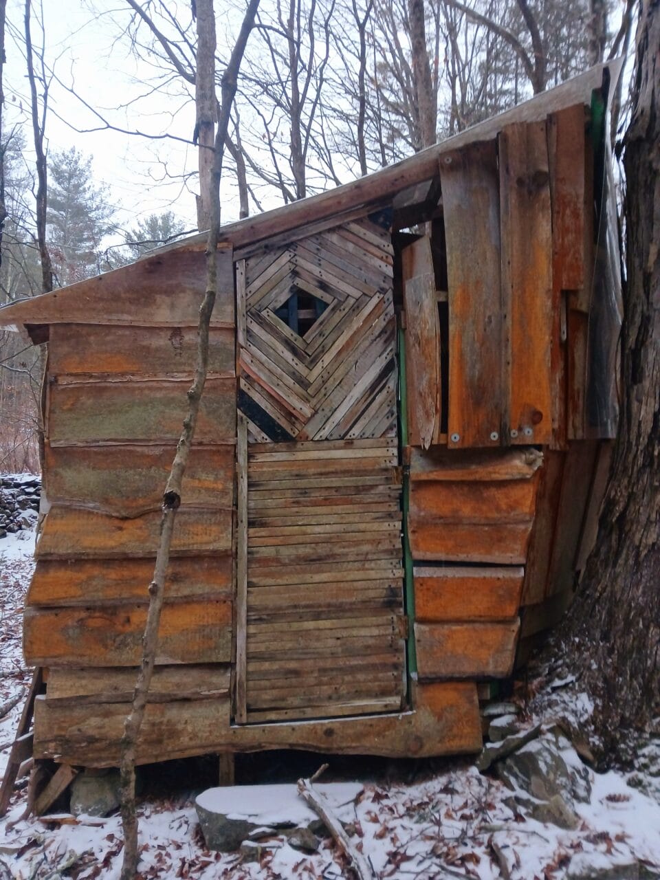 a hand-built shed-like structure in a snowy wooded landscape, part of "B-Home," a collection of intuitively built structures from salvaged materials