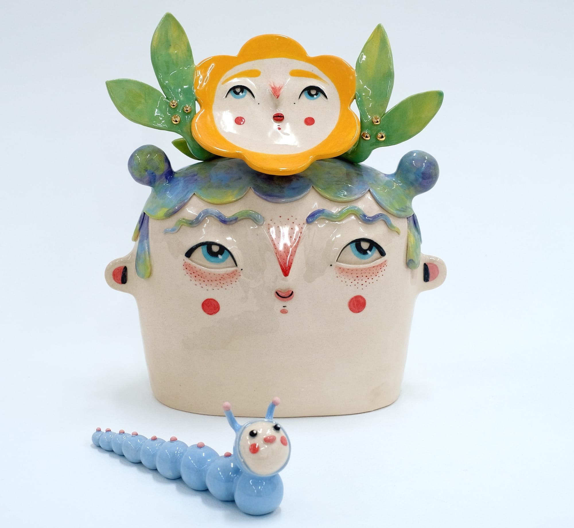 a ceramic sculpture of a face with a small flower with a face on its hair. a blue ceramic worm sits in front