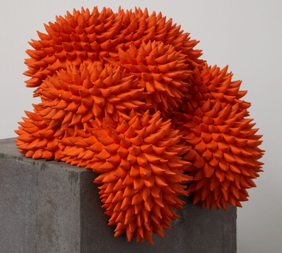 an amorphous orange sculpture with spines all over that appears to hang off a pedestal