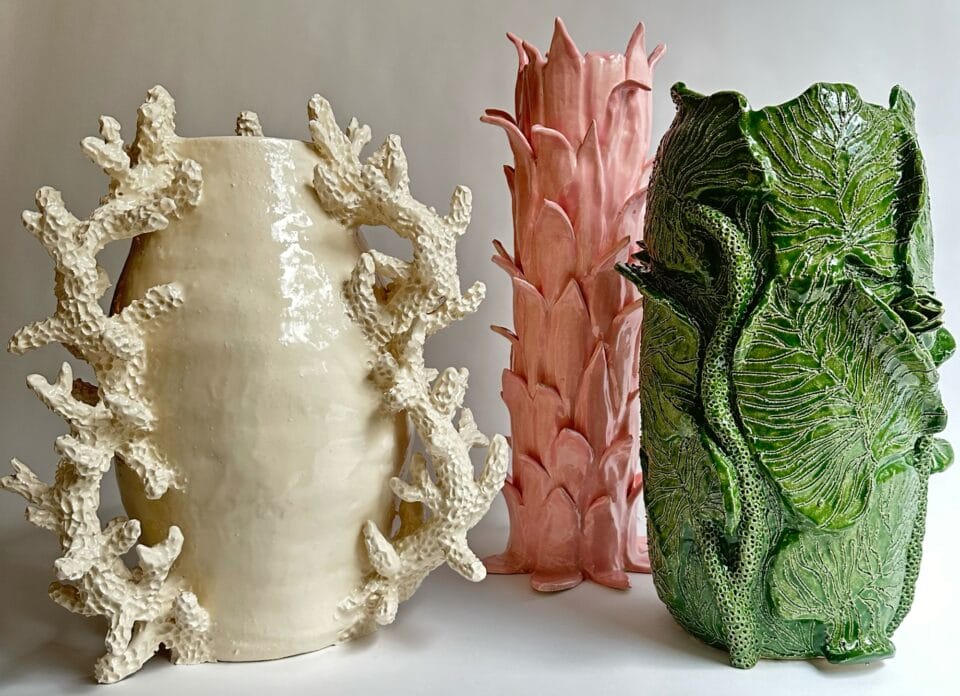 a trio of vessels, on the left is white with coral like handes,  the middle is pink with scalloped leaves, and the right is green with large cabbage like leaves