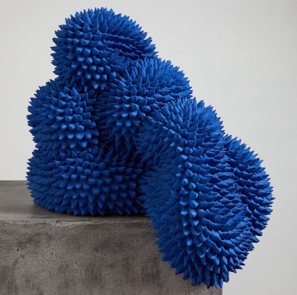 an amorphous blue sculpture with spines all over that appears to hang off a pedestal