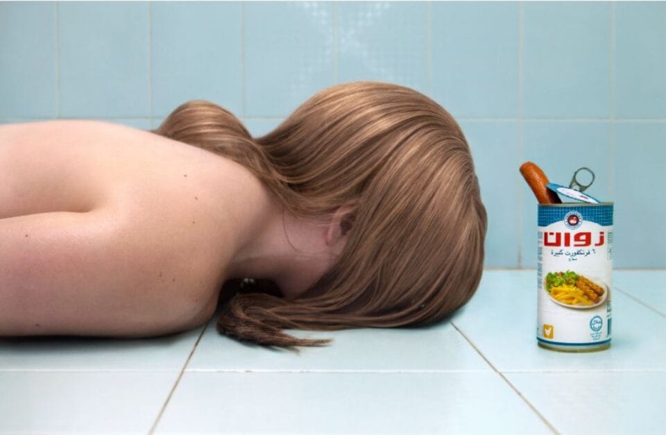 a photograph of a woman, face down on a tile floor, with her hair over her face and an open can of hot dogs next to her