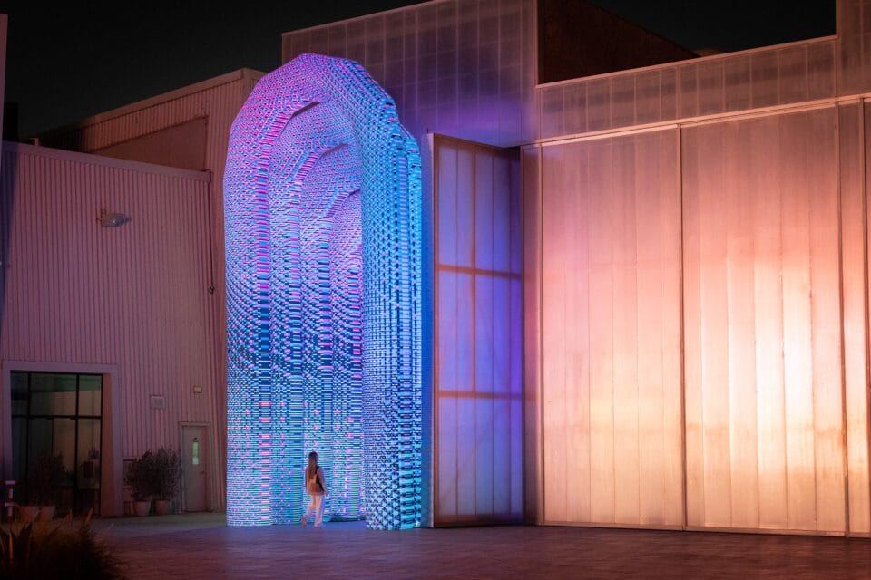 a monumental illuminated archway extending from a contemporary building with a person standing under the archway to show its large scale