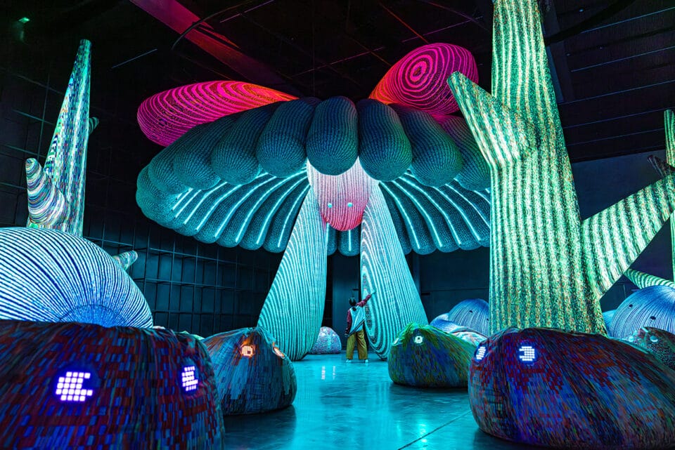 a person stands beneath an illuminated installation with lights resembling the underside of a mushroom