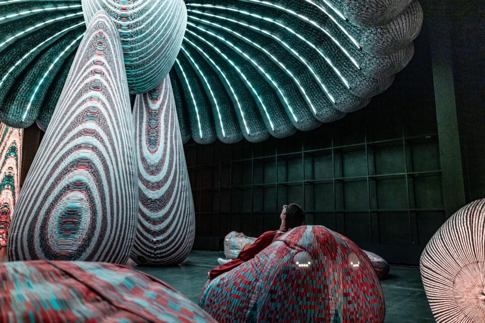 a person lounges in a beanbag-like seat beneath an illuminated installation with lights resembling the underside of a mushroom