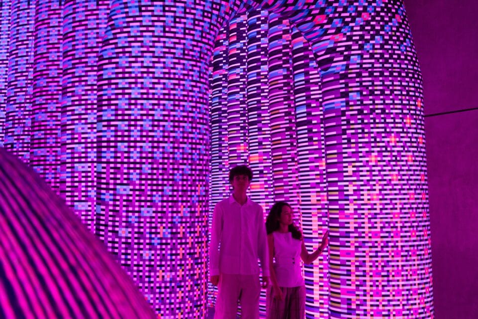 two people stand inside of an illuminated installation with a purple-and-pink geometric pattern