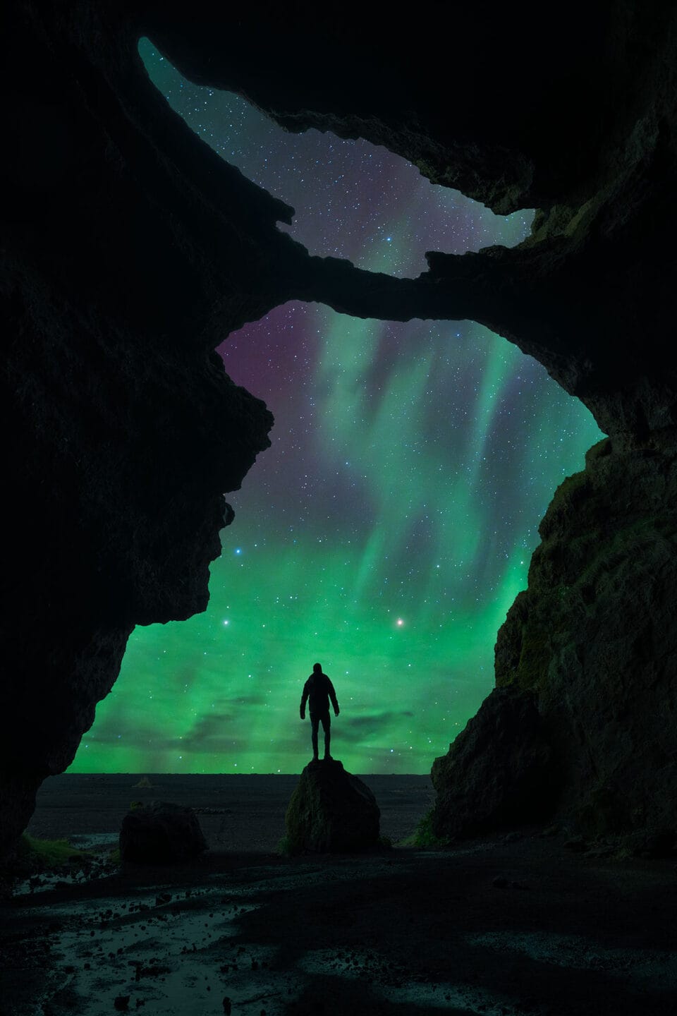 a figure stands in the opening of a cave, its silhouette accentuated against a glowing sky displaying the Northern lights.