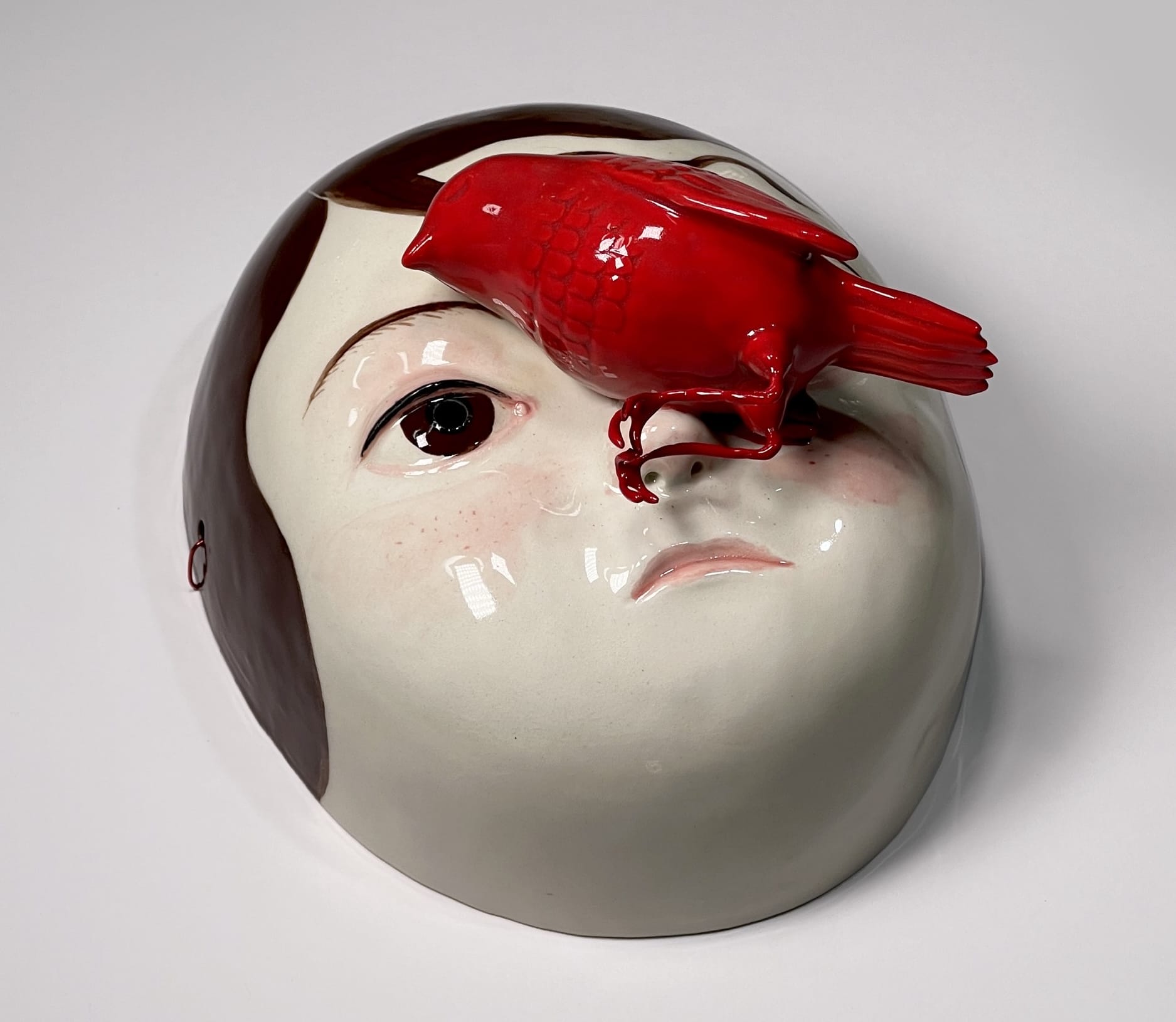 a ceramic mask with the face of a young girl with brown hair, with a dead red bird on her nose