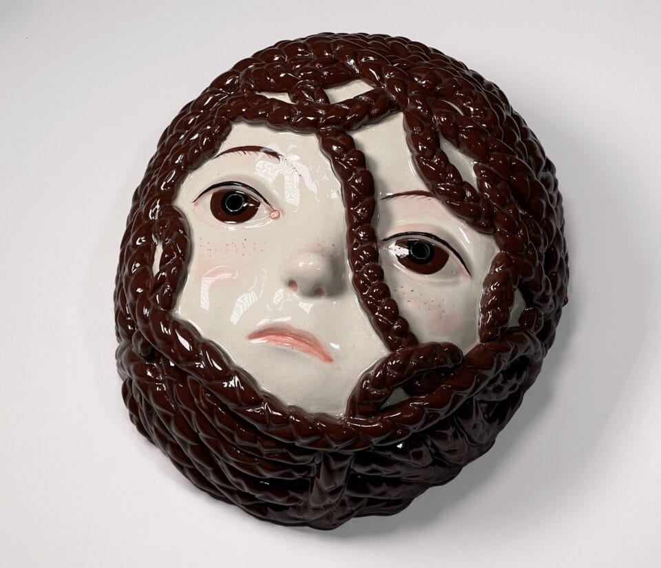 a ceramic mask with the face of a young girl with brown hair in braids that are engulfing her face