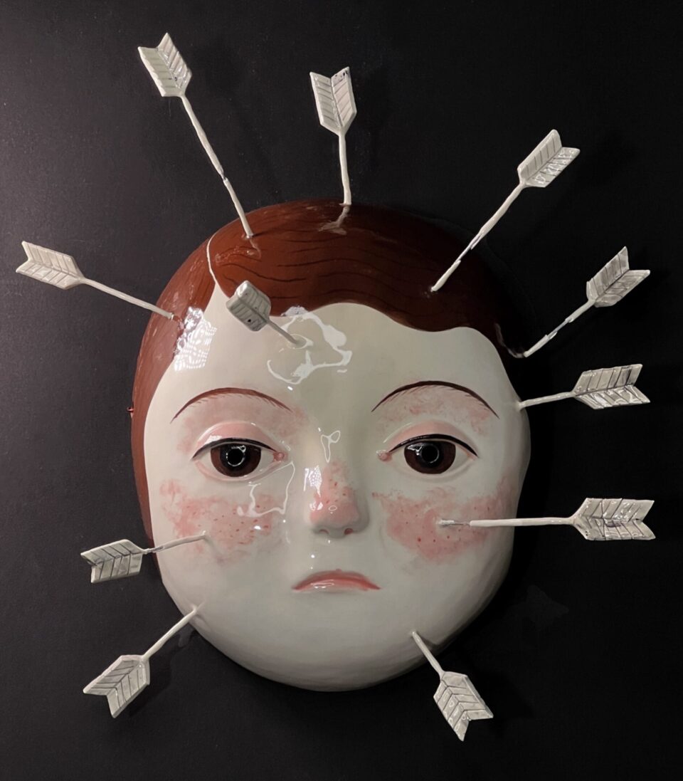 a ceramic mask with the face of a young girl with brown hair, with numerous arrows sticking out of her face