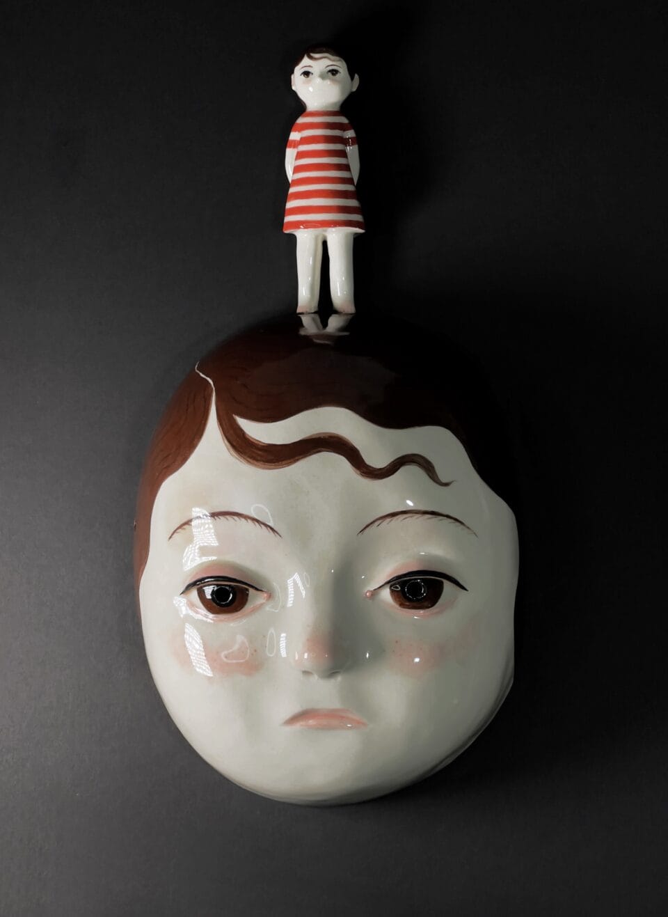 a ceramic mask with the face of a young girl with brown hair, with a small figure of another girl standing on the head, wearing a red-and-white striped dress