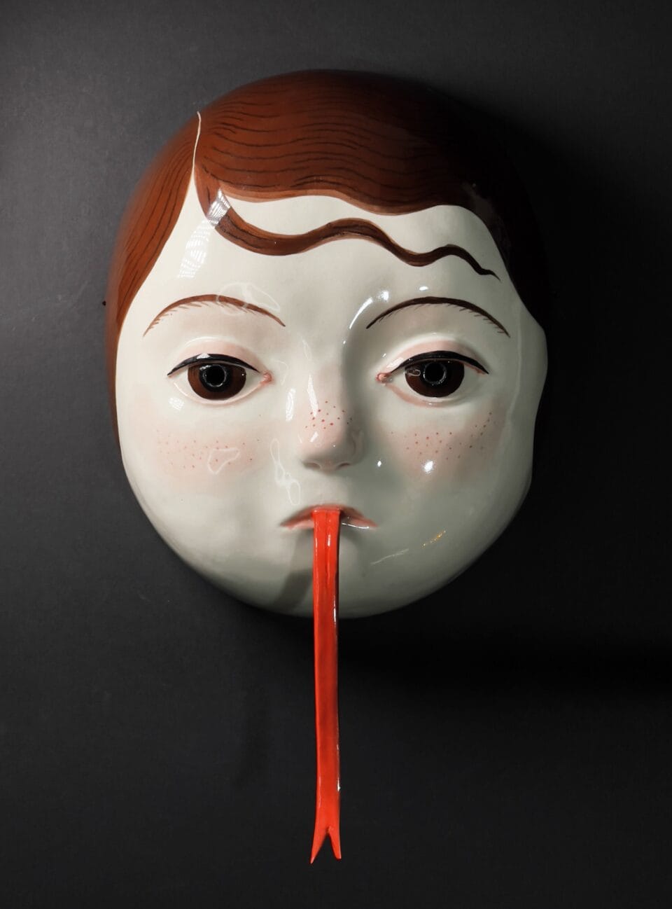 a ceramic mask with the face of a young girl with brown hair and a very long, skinny red tongue draped from her mouth