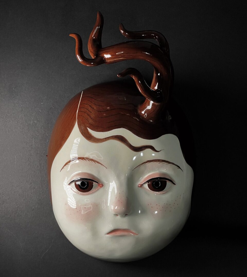 a ceramic mask with the face of a young girl whose hair is raising up in a flame-like tendril