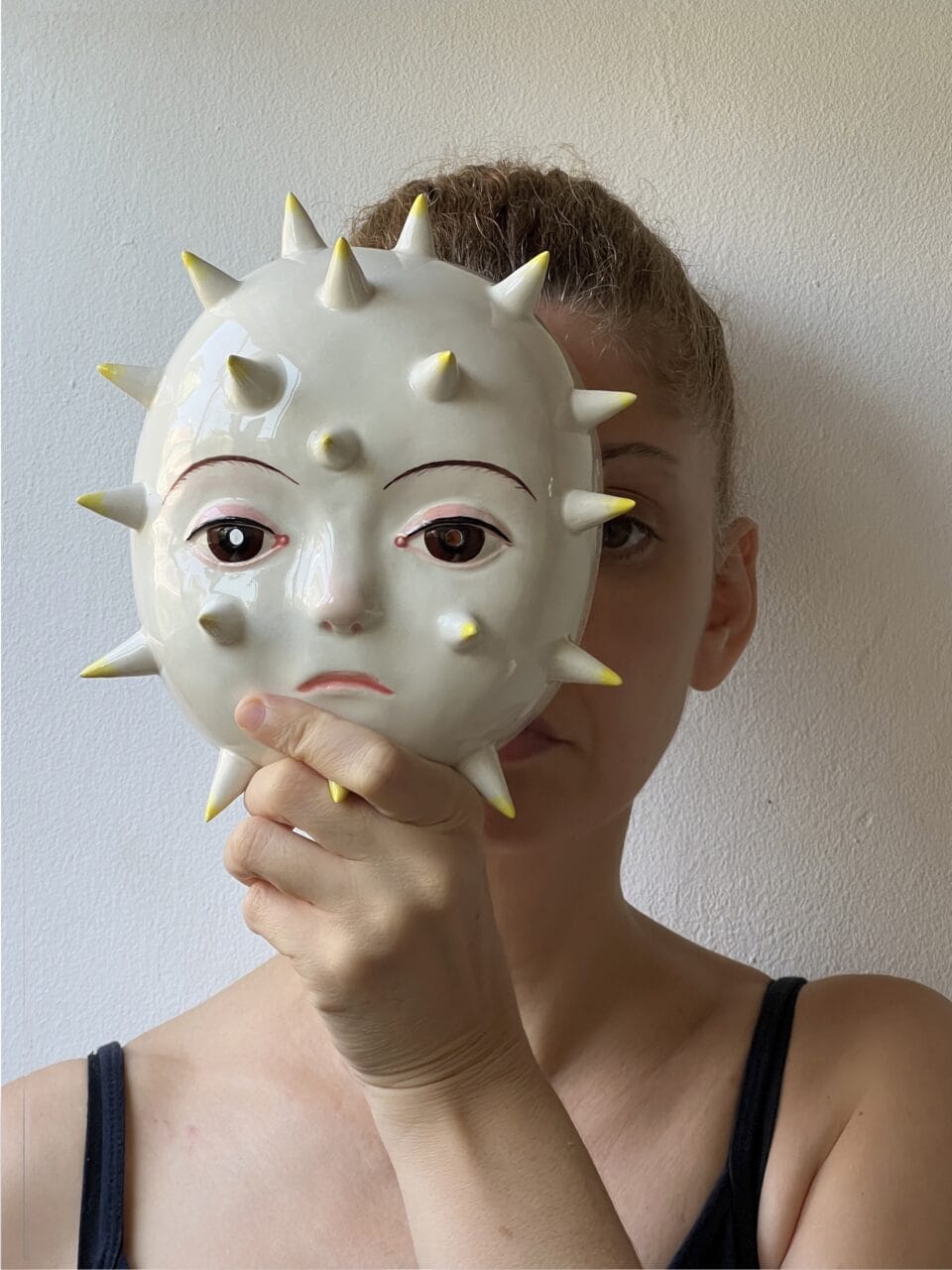 a figure holds a small ceramic mask in front of their face, featuring a white finish and spikes protruding from its surface