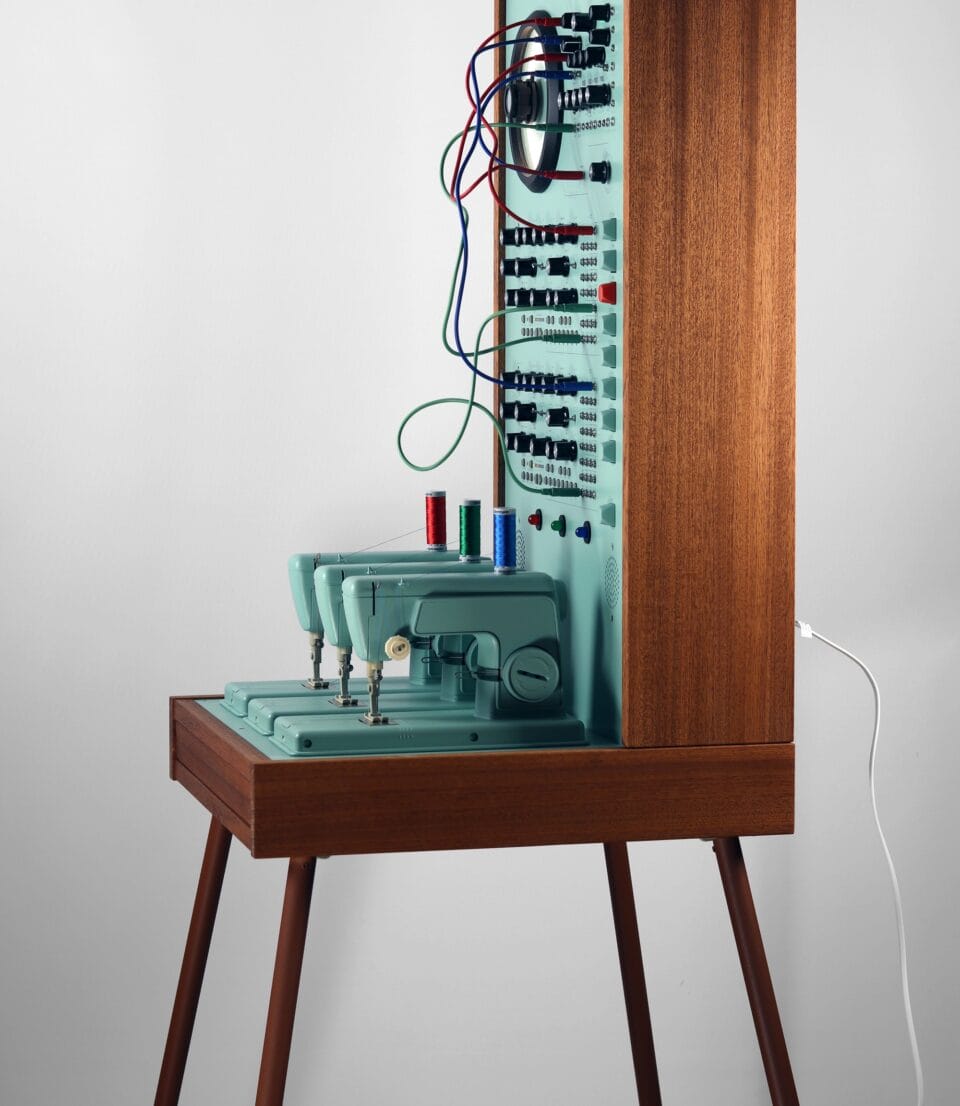 a sculpture resembling a retro computer or device in a teal hue with wood on the sides, with dials and wires and a row of three sewing machines on the base