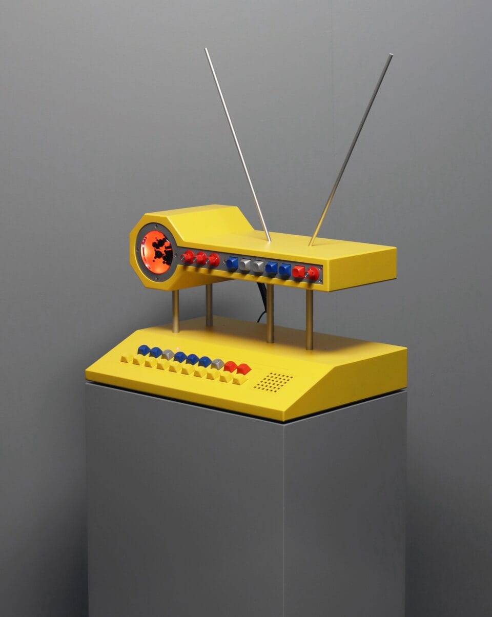 a sculpture resembling a retro computer or device in bright yellow with illuminated orange dials and black and white buttons