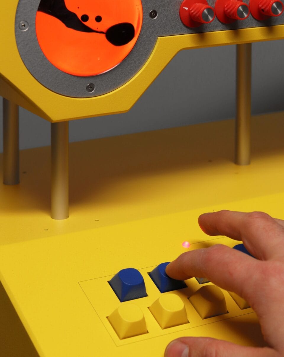 a detail of a sculpture resembling a retro computer or device in bright yellow, with a hand reaching out to press a black button