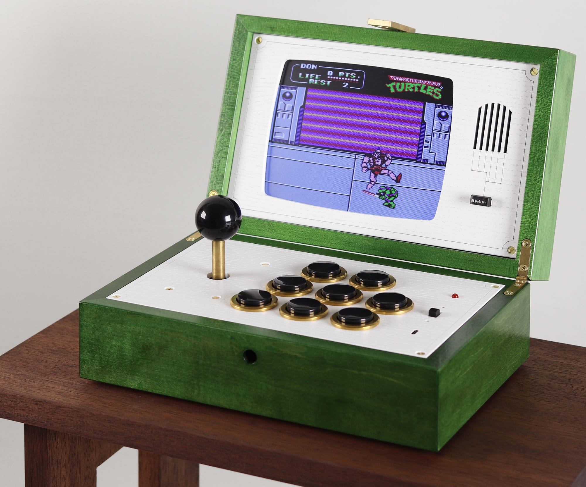 a sculpture resembling a retro, green computer or device with a hinged closure, shown open with a white surface, a black toggle stick and black buttons, and a small screen with a pixellated game image