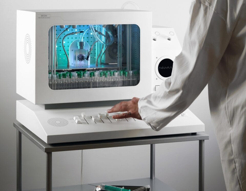 a sculpture resembling a retro computer or console in white, with a screen holding a bottle with tubing coming out of it and numerous tiny vials