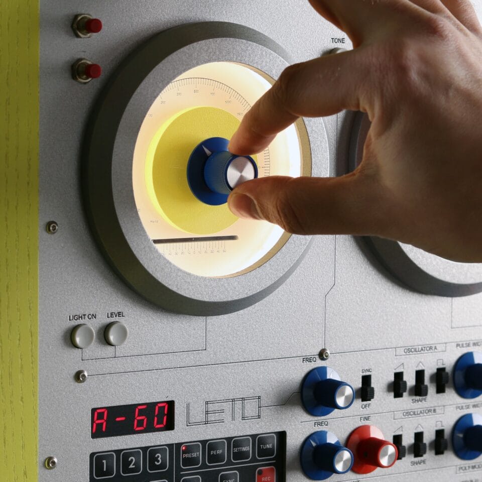 a detail of a sculpture resembling a retro computer or console in a bright yellow hue with a silver backing for numerous black buttons and dials, shown with a hand adjusting a knob