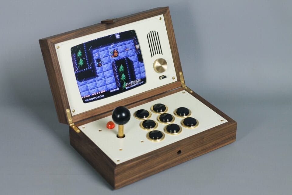 a sculpture resembling a retro computer or device with a hinged closure, shown open with a white surface, a black toggle stick and black buttons, and a small screen with a pixellated game image