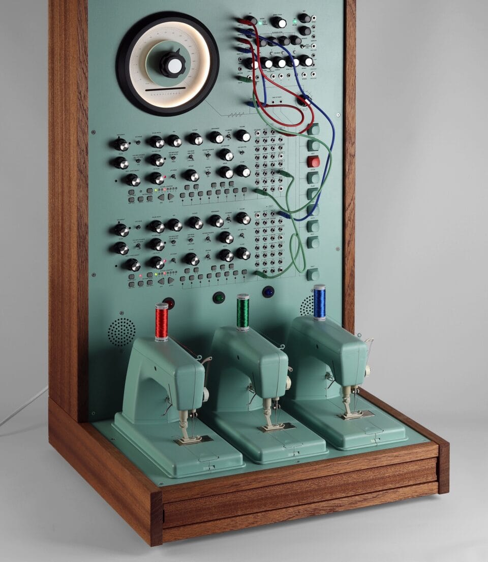 a sculpture resembling a retro computer or device in a teal hue with wood on the sides, with dials and wires and a row of three sewing machines on the base