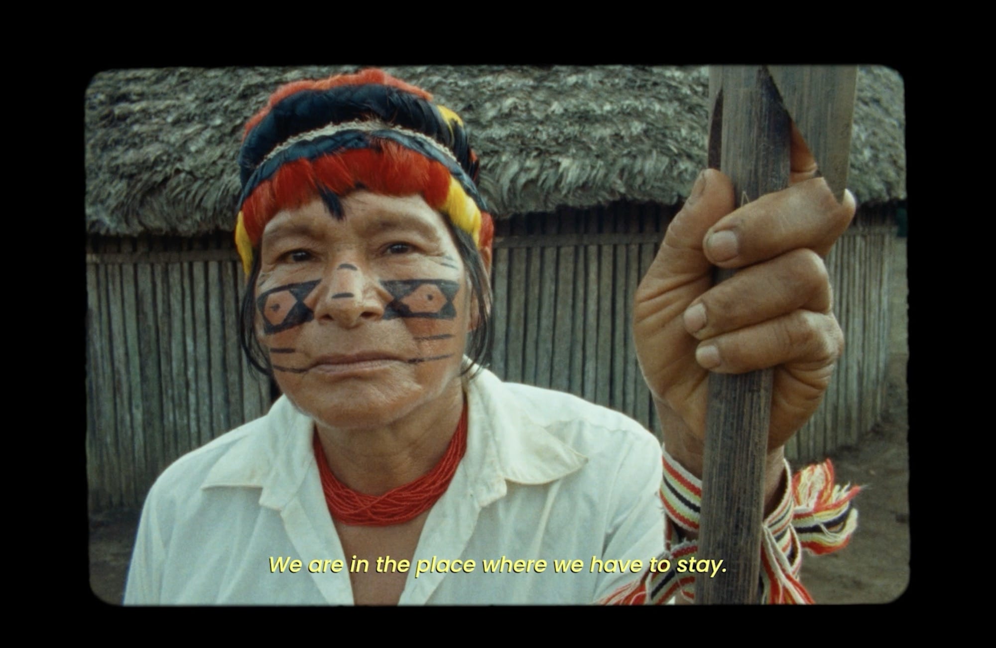 a still from the short documentary film 'I Am the Nature' featuring an Indigenous Achuar woman with traditional face paint and textile headband, with the subtitle text reading "We are in the place that we have to be"