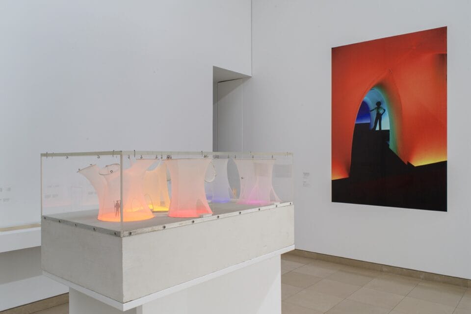 an installation view of a photograph of a colorful fabric installation and a small case with a maquette of a large-scale, immersive textile environment