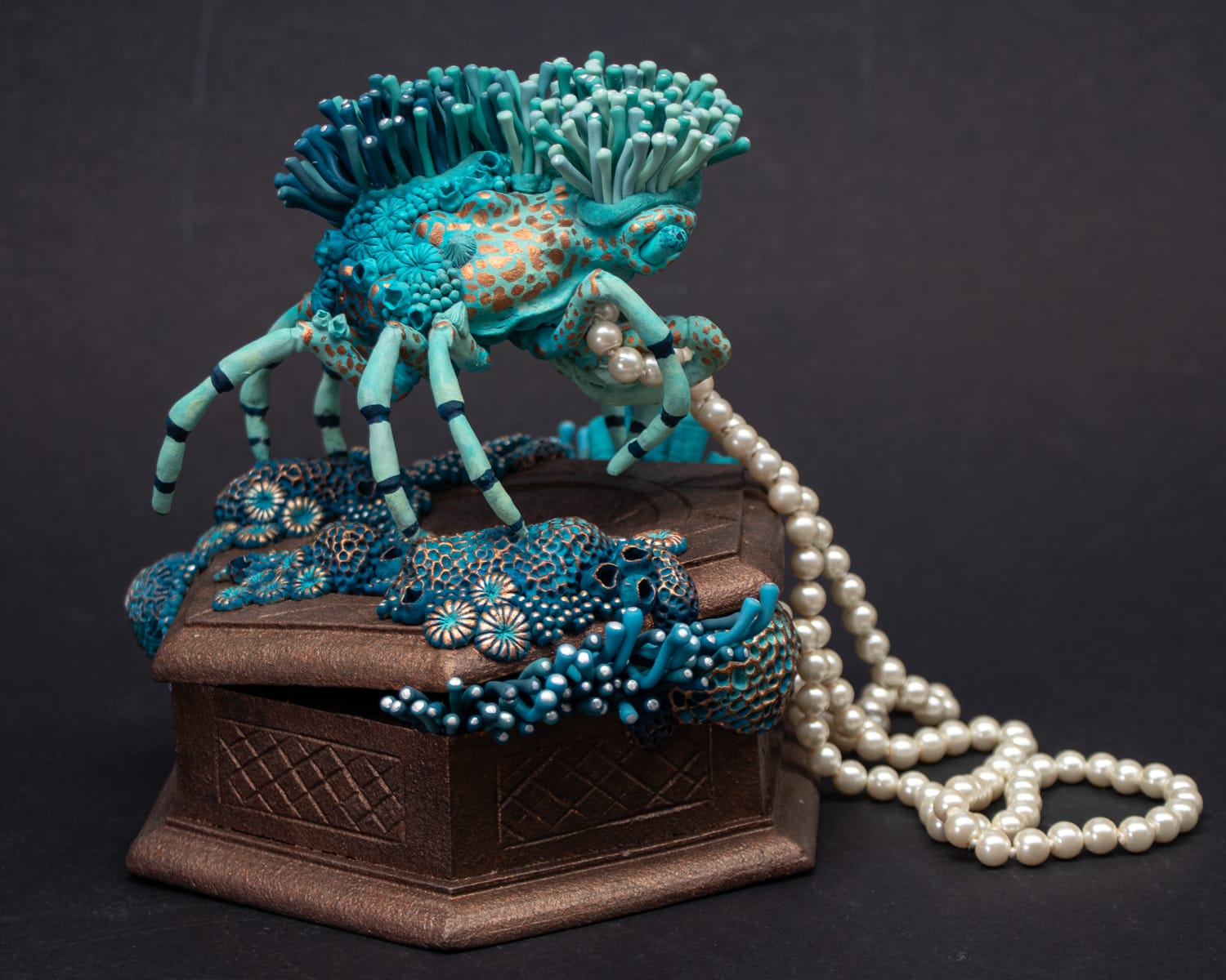 a sculpture made from a vintage box with a decorator crab on top, collecting a string of pearls and surrounded by blue coral