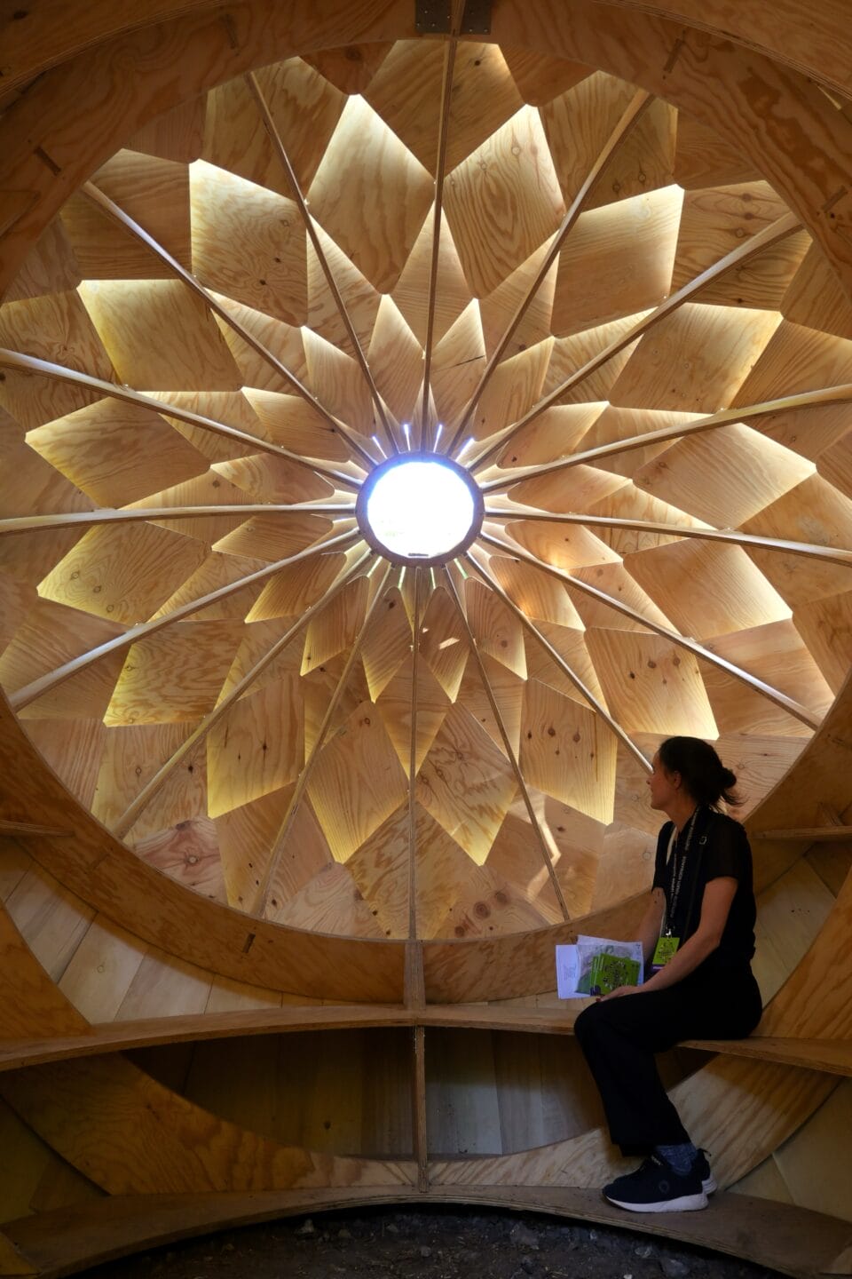 a woman in black clothing sits inside a wooden structure on a bench. sun streams through a round opening surrounded by layered diamond shapes