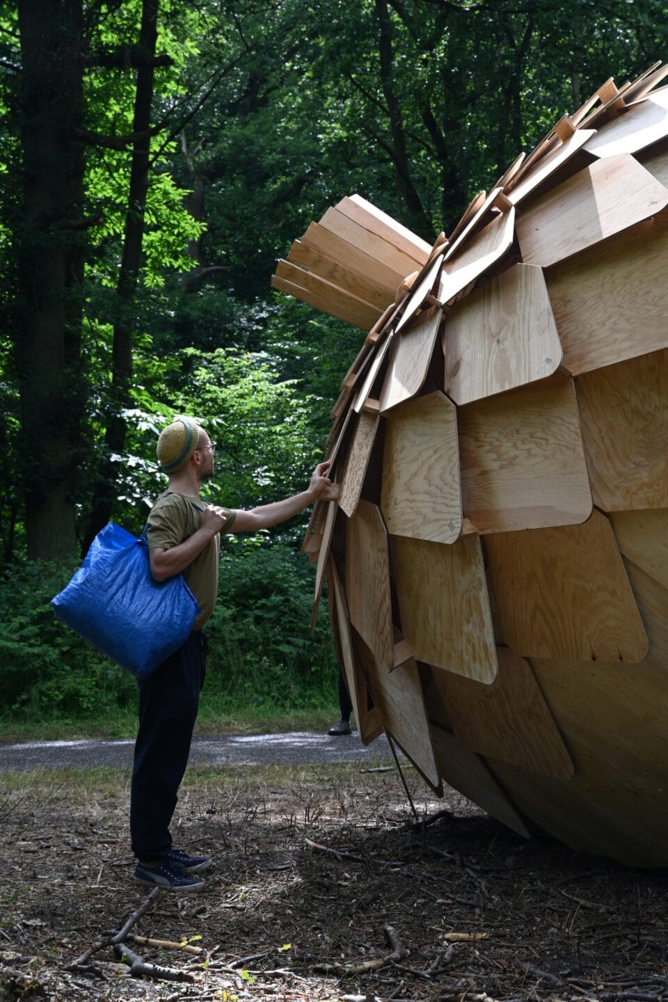 a person holding a blue bag touches part of a large wooden acorn resting in a forest