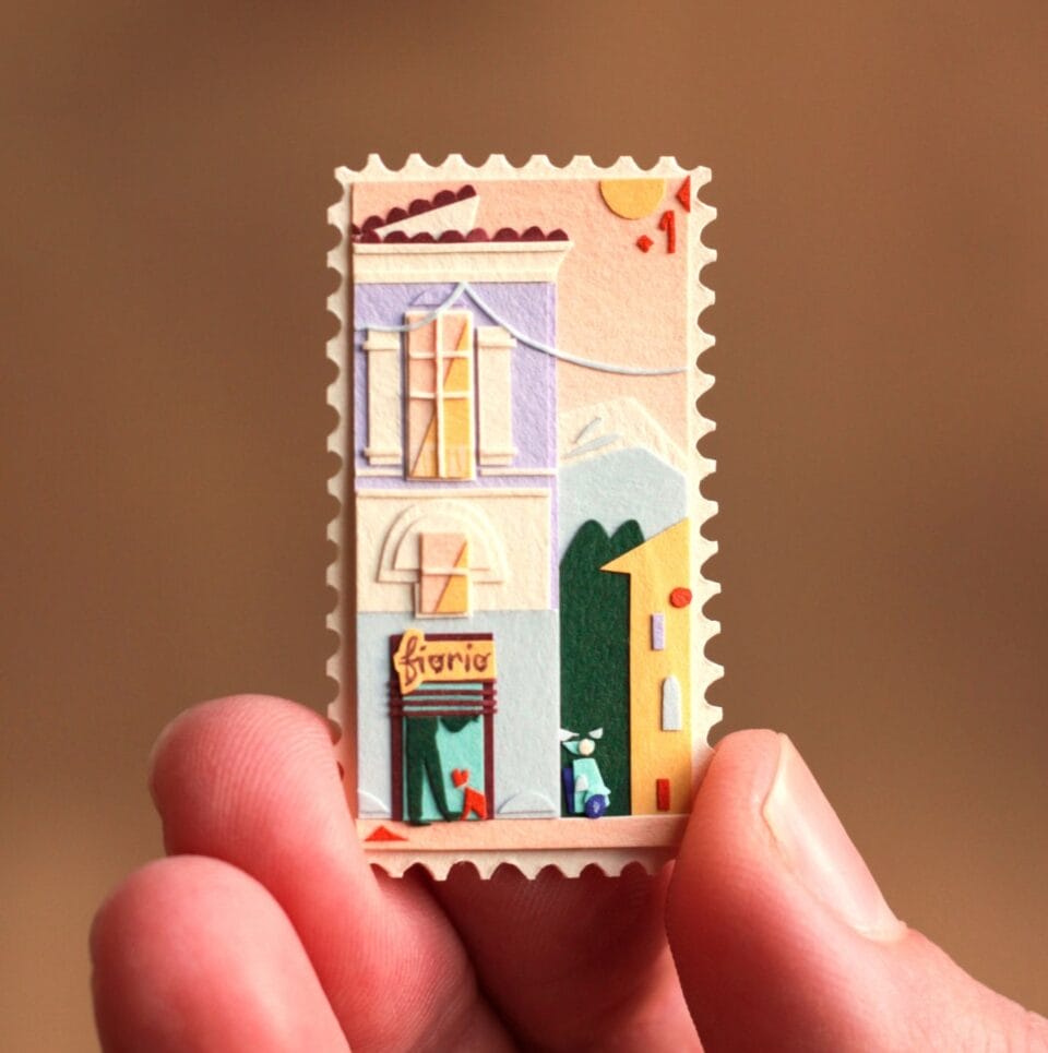 a hand holds up a small, vibrant collage in the shape of a stamp. it depicts a scene with a building and patterned designs