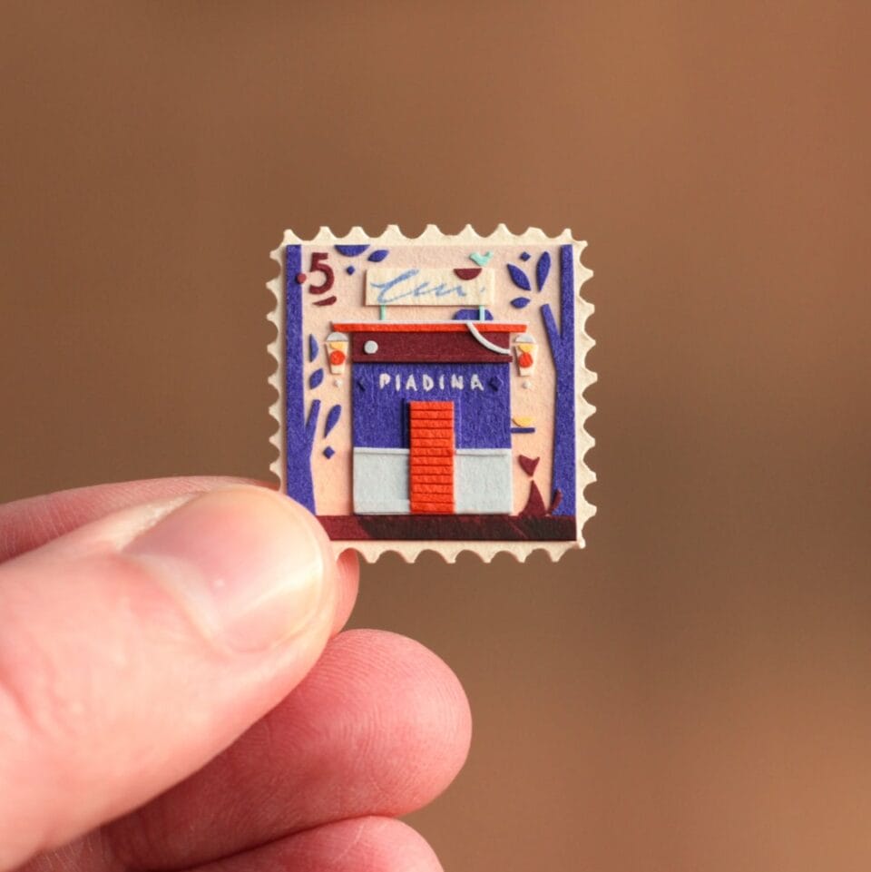 a hand holds up a small, vibrant collage in the shape of a stamp. it depicts a scene with a building and patterned designs