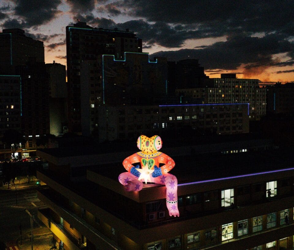 an aerial view of an inflatable public installation of a fantastical, brightly-colored figure perched on the edge of a city building, illuminated at night