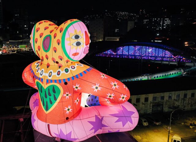 an aerial view of the rear of an inflatable public installation of a fantastical, brightly-colored figure perched on the edge of a city building, illuminated at night