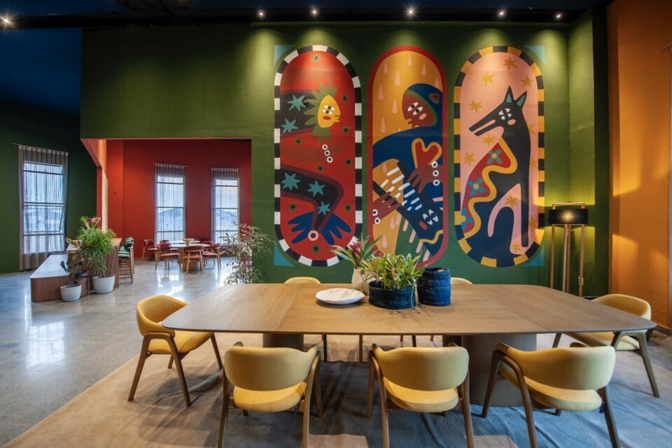 a mural on an interior wall of a gathering space with tables and chairs in the foreground. the mural has three panels showing a fantastical fish, a person, and a wolf