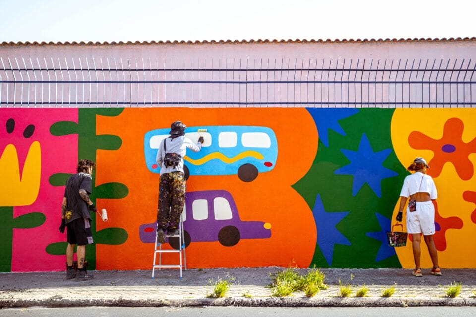 artists working on a long, colorful mural on the side of a building