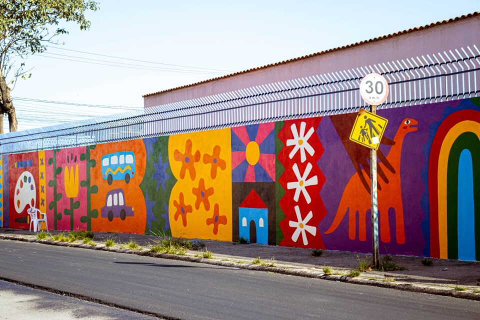 a long mural on the side of a school building in Brazil depicting flowers, a painting palette, a red dinosaur, a rainbow, and other joyful motifs