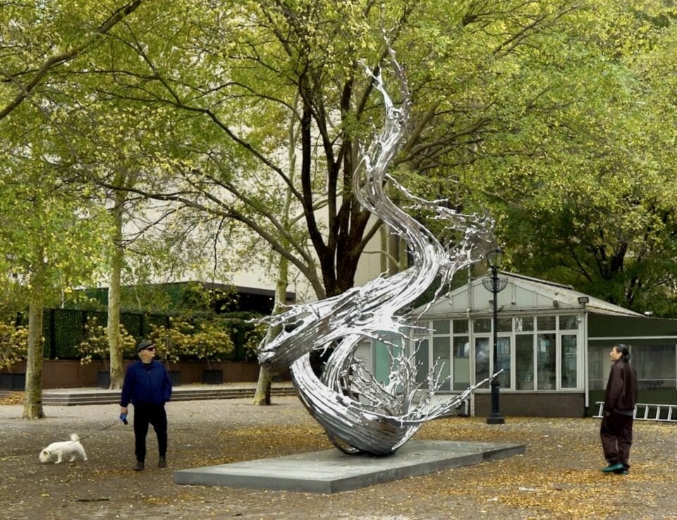 a chrome-colored abstract sculpture installed in a public park that resembles a large swirling splash of liquid that has been frozen in time